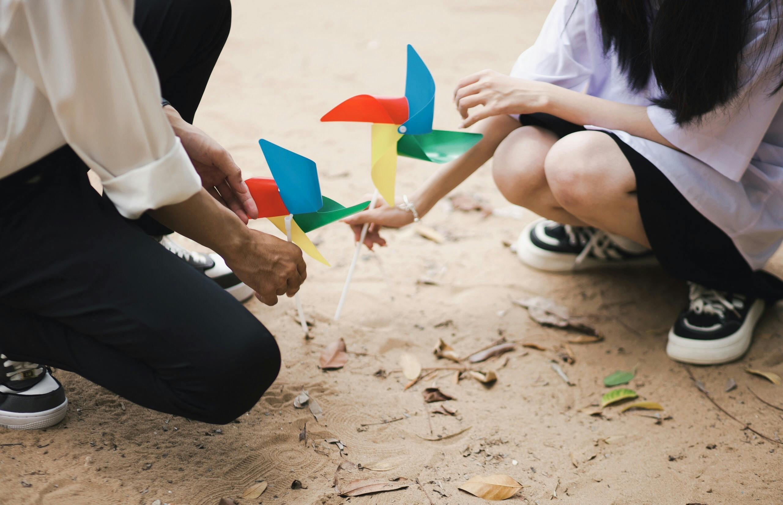 Adults kneeling on sand, engaging with colorful pinwheels, symbolizing play and creativity.